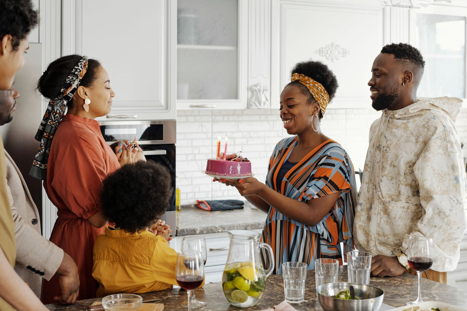 A family gathered in a kitchen as a woman holds a birthday cake with lit candles