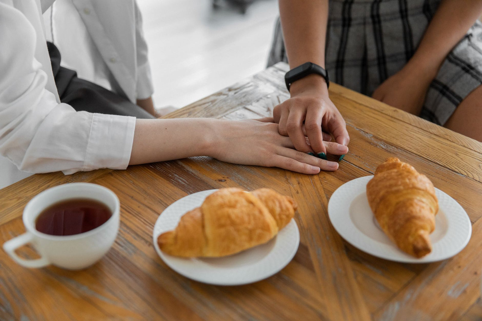 A married couple resting their hands together across a wooden kitchen table over coffee and pastries