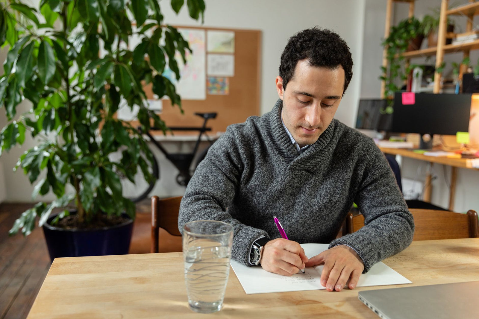 A man in a grey sweater writing by hand at a wooden desk